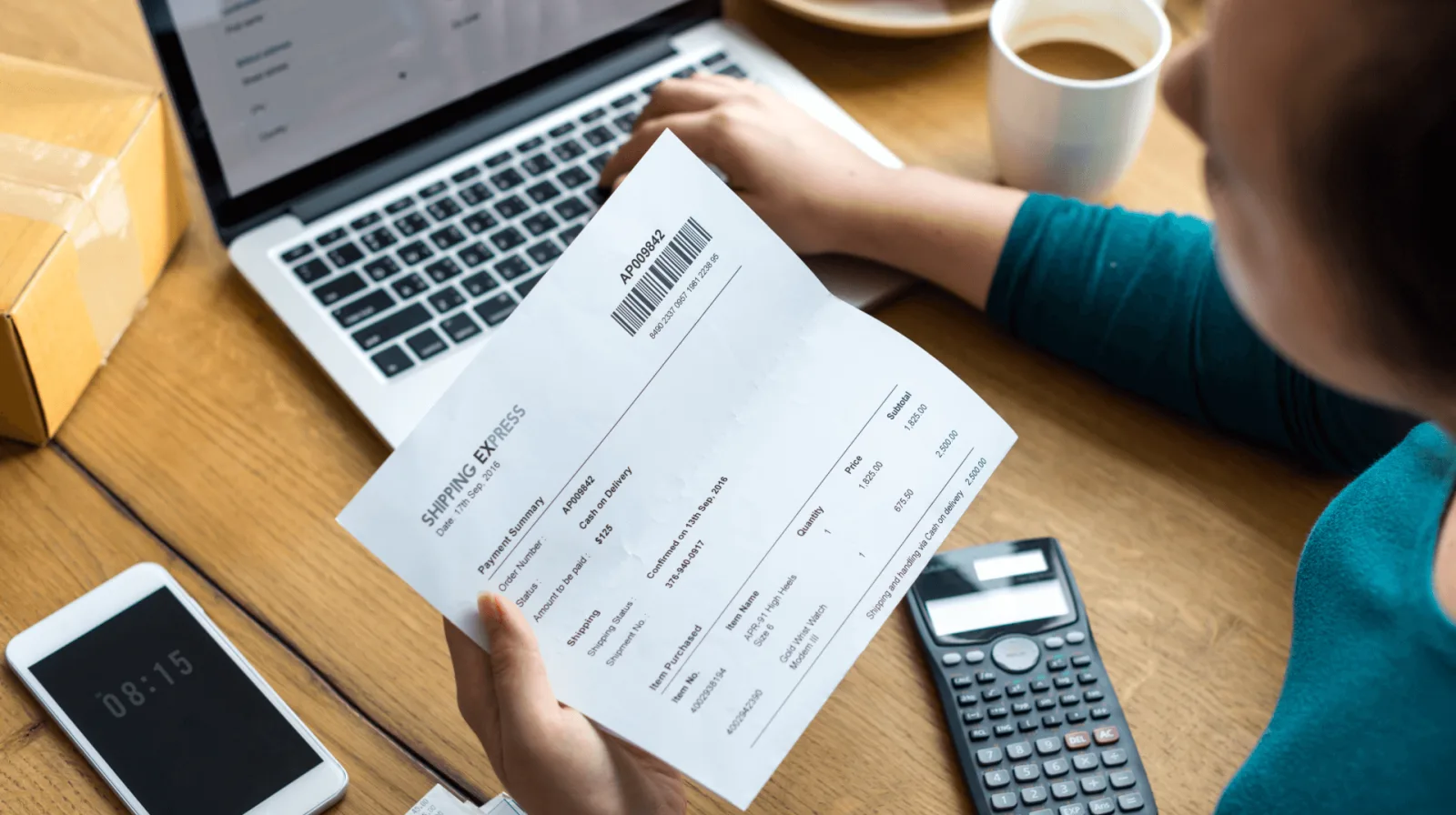 A person holding a printed shipping document at a desk with a laptop, a calculator and a phone