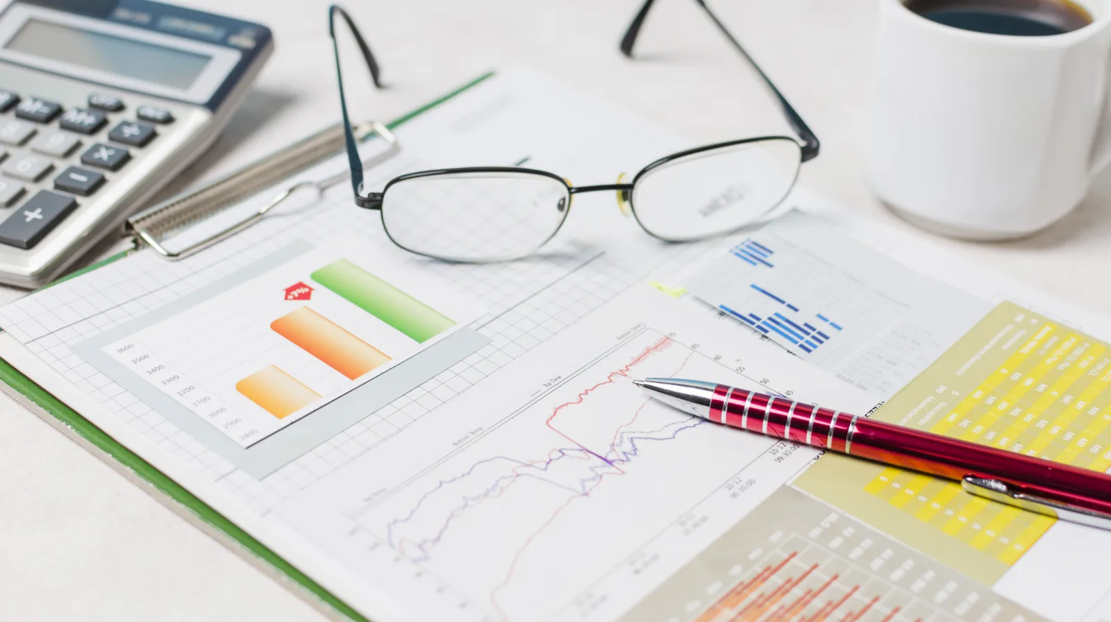 Printed financial reports with bar and line charts on a white desk, beside reading glasses, a pen, a calculator and a coffee cup