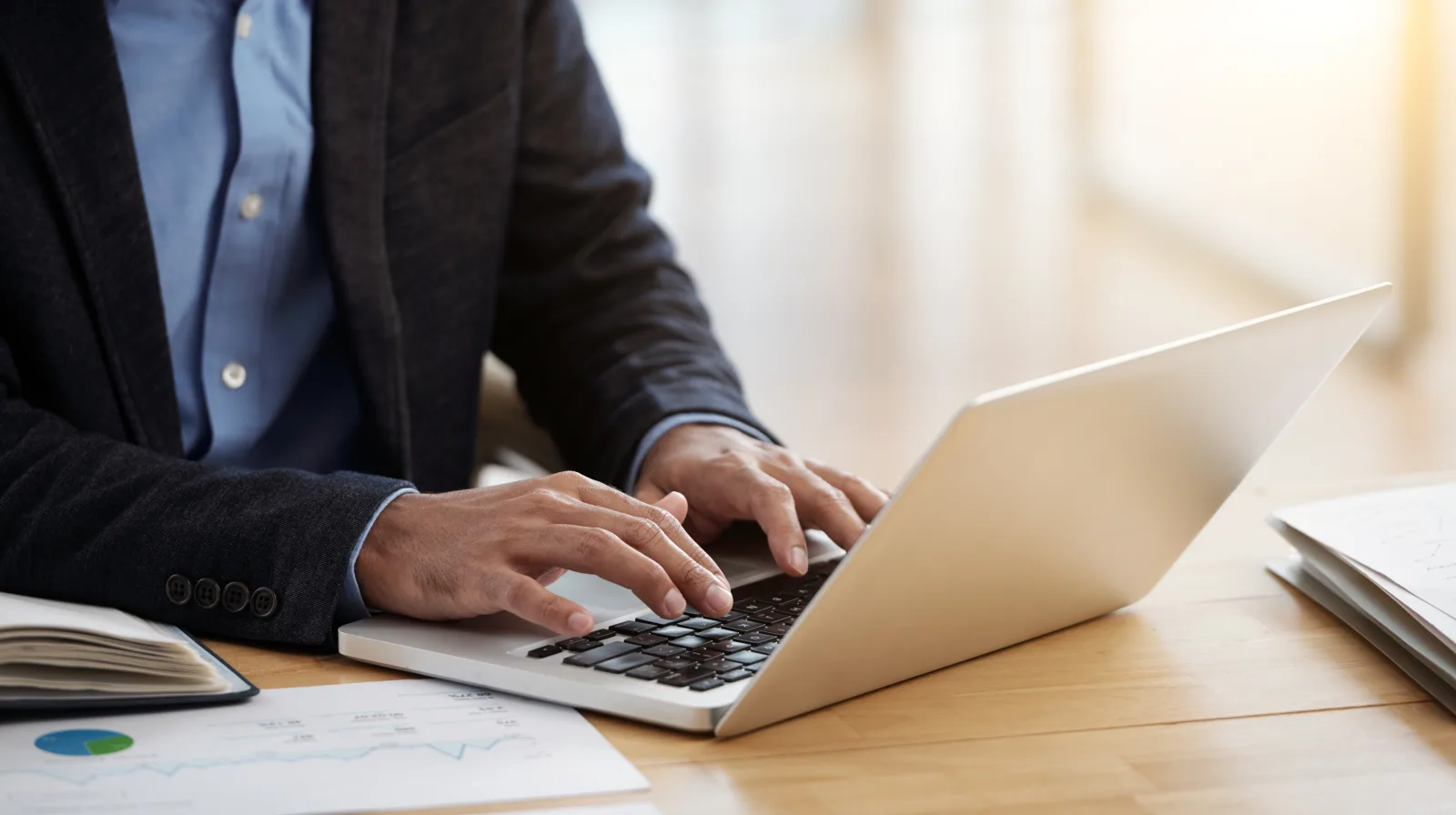 A person typing on a laptop at a bright desk, with printed charts and a notebook beside it