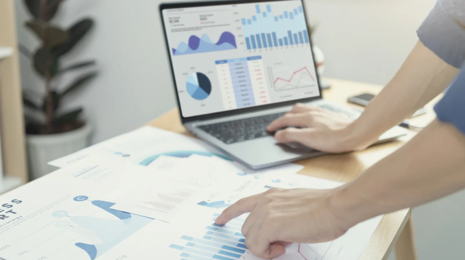 Hands at a laptop showing charts, beside printed business reports on a bright desk