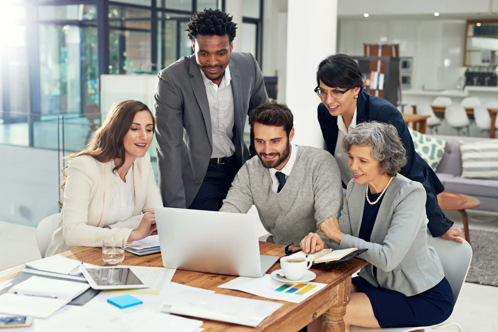 A finance team gathered around a laptop at a table covered with printed reports, in a bright office